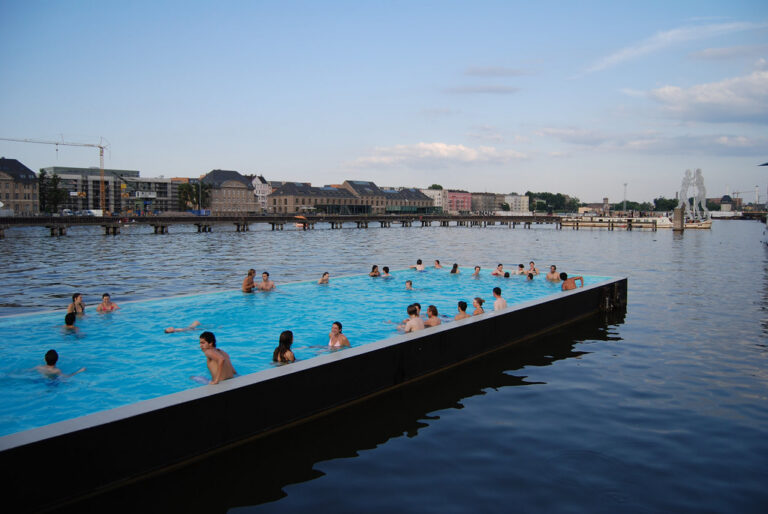 People swimming in a bright blue outdoor pool on a river on a sunny day