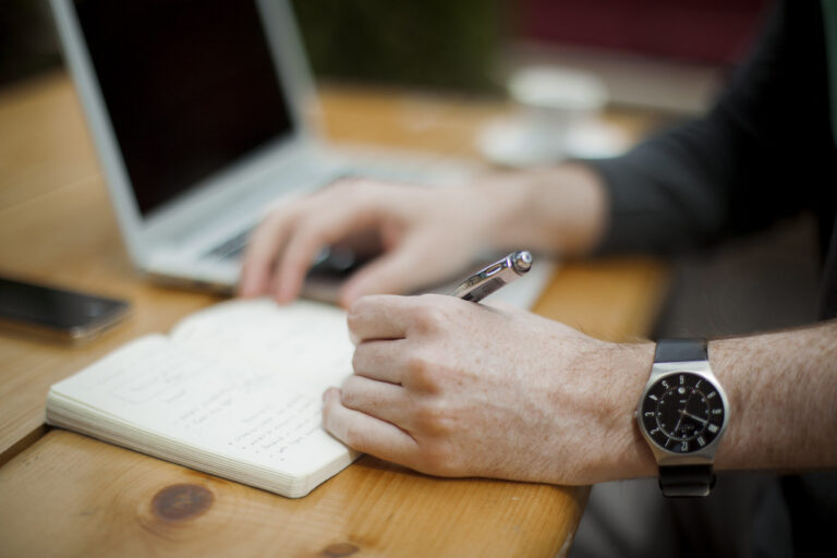 Hand writing in an open notebook at a wooden desk beside a laptop