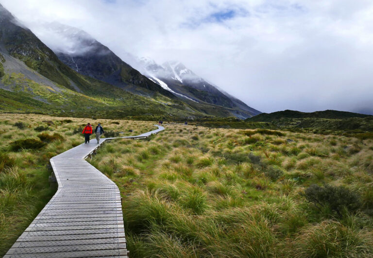 People walking along a wooden boardwalk through green valley grassland toward mountains