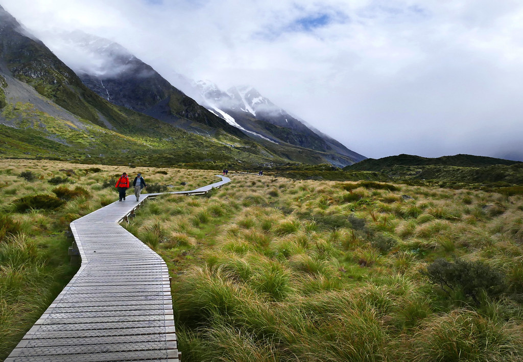 People walking along a wooden boardwalk through green valley grassland toward mountains