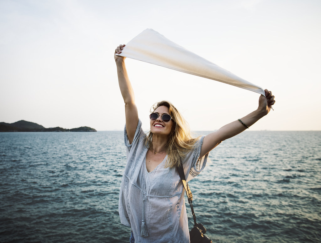Smiling woman wearing sunglasses holding a light scarf in the breeze beside the sea