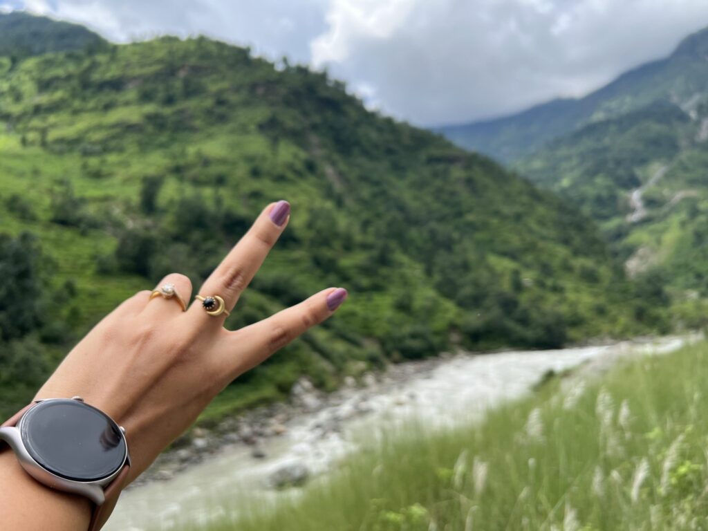 Hand wearing a smartwatch making a peace sign above a green mountain valley