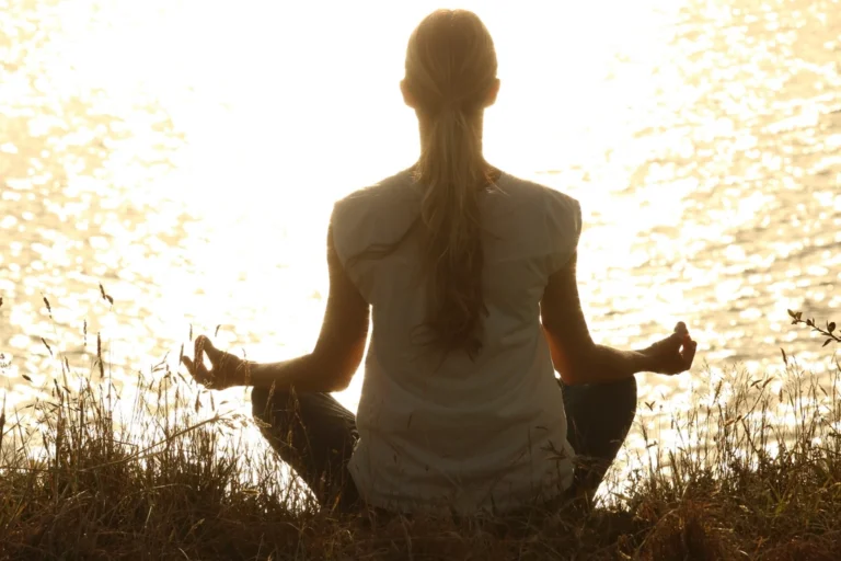 Woman sitting cross-legged and meditating by sunlit water on a bright day