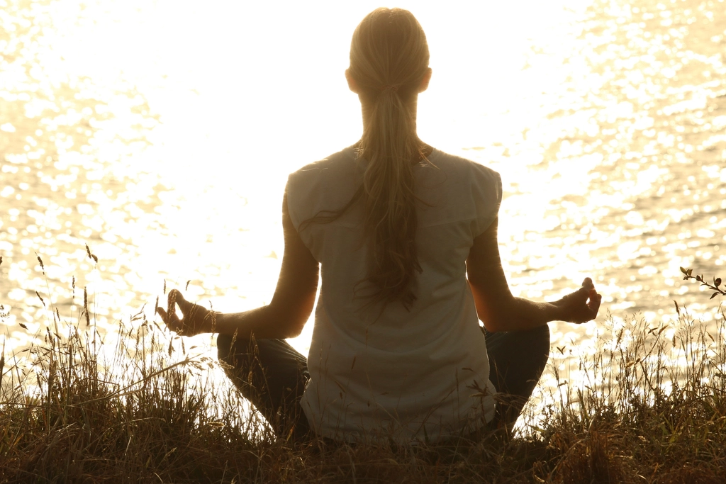 Woman sitting cross-legged and meditating by sunlit water on a bright day