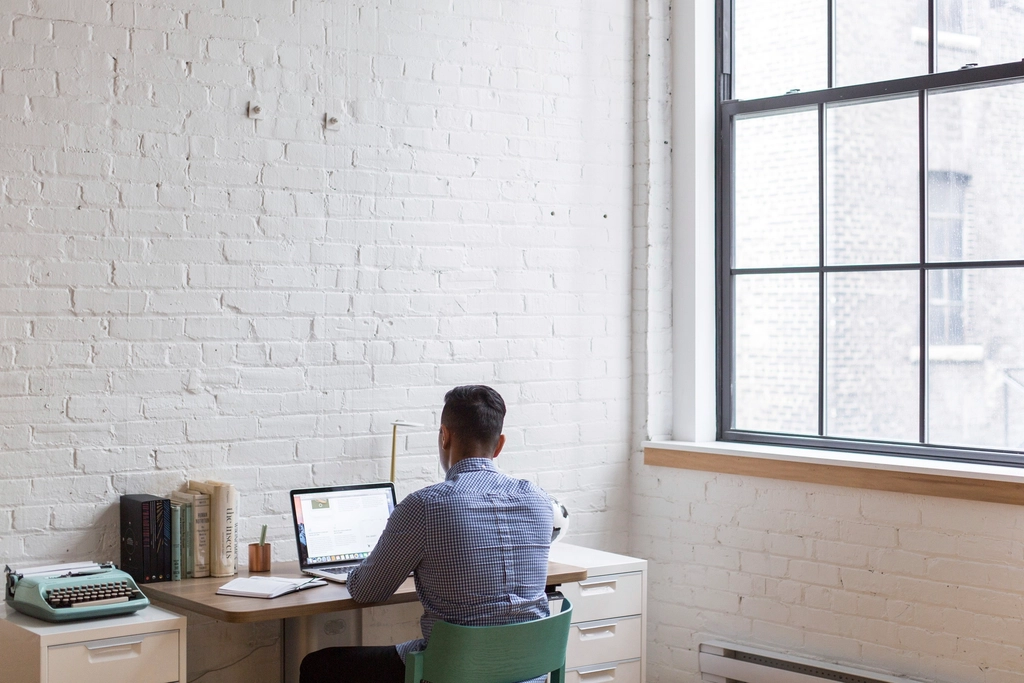 Man working on a laptop at a tidy desk beside a large bright window