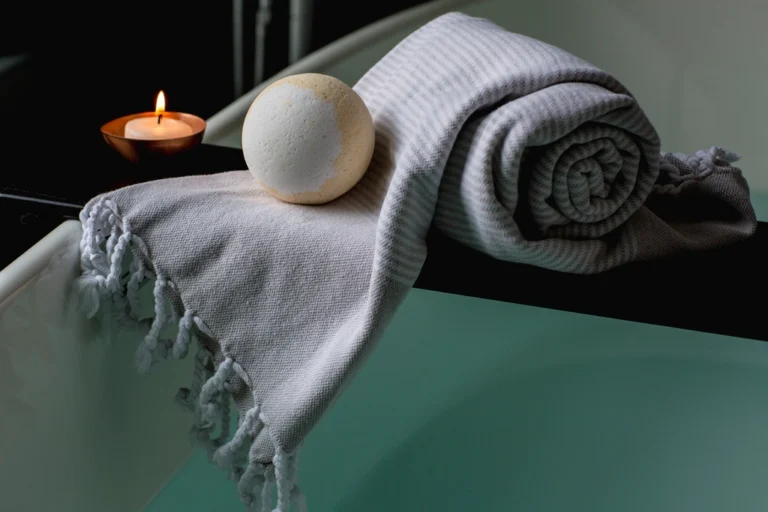 Rolled bath towel, bath bomb and a lit candle on the edge of a bathtub