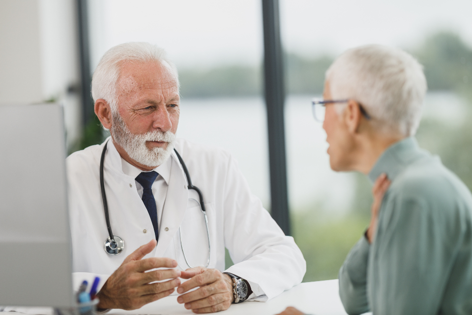 Senior doctor in a white coat listening to an older woman patient at his desk