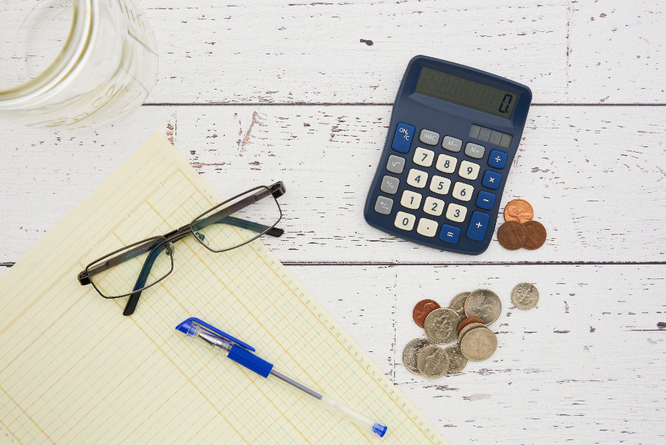 Calculator, glasses, pen, coins and a notepad on a white wooden desk